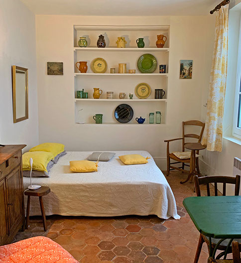 Studio interior with shelves of pottery, queen bed, mirror, armchair and small oval table, green dining table, bistro chairs.